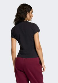 Čierne tričko s krátkym rukávom vyrobené z bavlny, priliehavý strih, s hladkou textúrou. Nosené s vínovými teplákmi, zobrazené zozadu.