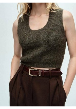Kobieta ubrana w oliwkowy, dziergany crop top bez rękawów, zestawiony z ciemnobrązowymi, wysokimi spodniami i ciemnobrązowym paskiem.