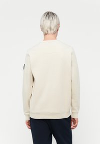 Light beige sweatshirt with a crew neck, long sleeves, ribbed cuffs and hem, featuring a small back patch detail. Worn over dark pants.