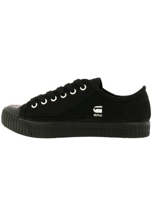 Zapatilla negra de lona de caña baja con suela de goma negra, cordones negros, ojales metálicos y un pequeño logo blanco "RAW" en el lateral.