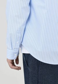 Chemise rayée bleu clair avec un ourlet incurvé, poignet boutonné et texture douce, associée à un jean rayé foncé, montrant une main près de la poche.
