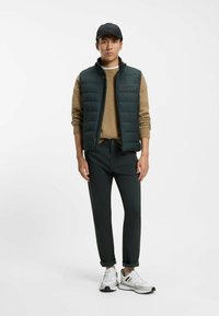 Gilet imbottito verde scuro sopra un maglione color sabbia, abbinato a pantaloni slim fit verde scuro e sneakers bianche con dettagli arancioni e grigi.