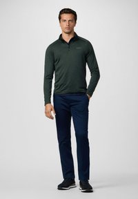 Dunkelgrüner Langarm-Pullover mit Kragen und Reißverschluss, kombiniert mit marineblauen Hosen und schwarzen Sneakern. Der Pullover hat ein dezentes Logo auf der Brust.