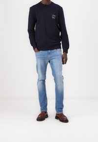 Uomo che indossa un felpa blu navy con logo, jeans azzurri e mocassini in pelle marrone, in piedi davanti a uno sfondo bianco semplice.