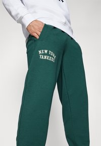 Zaļas sporta bikses ar mīkstu materiālu, kurām ir sānu kabatas un uz priekšpuses ir balti burti "NEW YORK YANKEES". Ikdienišķs sašaurināts dizains.