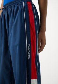 Marine treningsbukser med røde og hvite side-striper, lett stoff, elastisk midje, brodert "TOMMY JEANS"-logo på benet.