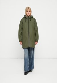 Grønn, hettejakke med vanntett materiale, glidelås, elastiske mansjetter og frontlommer, sammen med brede, blå denimjeans.