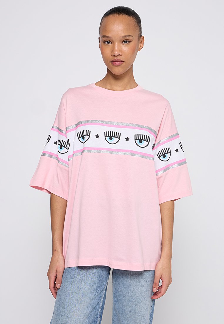 T-shirt en coton rose avec des manches courtes et larges, présentant une bande horizontale d'yeux graphiques et d'étoiles, rehaussée de rayures argentées et roses.
