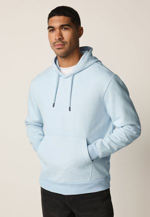 Muž nosící světle modrou mikinu s rukama v přední kapse, stojící před jednoduchým béžovým pozadím.