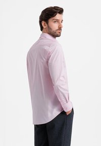 Camisa de manga larga rosa claro con cuello puntiagudo, puños abotonados y dobladillo curvado, combinada con pantalones gris oscuro.