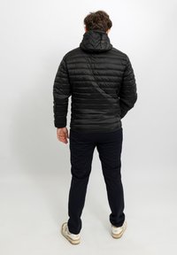 Giacca a vento nera con motivo trapuntato orizzontale, cappuccio attaccato e finitura lucida, indossata con pantaloni blu navy e sneakers bianche.