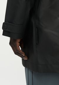 Δεξί χέρι ενός ατόμου που φοράει μαύρο παλτό με κουμπωμένο μανίκι, ακουμπισμένο δίπλα σε μπλε πλισέ παντελόνι με απλό φόντο.