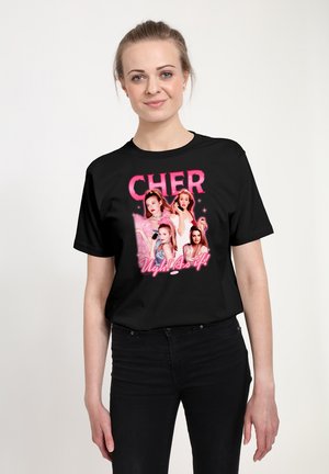 Kobieta o jasnej karnacji i związanych do tyłu włosach nosi czarną koszulkę z czterema zdjęciami Cher i różowym napisem "CHER" oraz "Ugh! As if!"