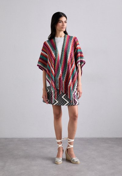 Kimono tejido multicolor con patrón en zigzag, adornado con flecos, combinado con una falda en chevron en blanco y negro y zapatos de esparto con tiras.