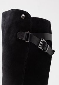 Botte haute jusqu'au genou en daim noir avec un dessus arrondi, équipée d'une bride en cuir noir et d'une boucle argentée sur le côté. Texture lisse.