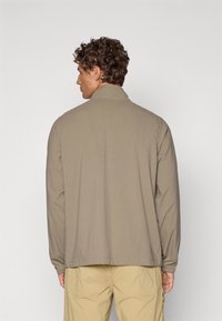 Jachetă ușoară de culoare khaki, cu guler înălțat și mâneci lungi, având o textură netedă și detalii minime de cusătură pe spate.