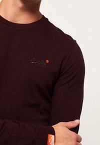 Burgundy pitkähihaisessa T-paidassa on tekstuuri, jossa on pieni musta logo rinnassa ja oranssi merkki hihassa.