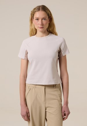Donna bionda che indossa una maglietta a maniche corte color beige chiaro con pannelli laterali più scuri e pantaloni beige, in piedi davanti a uno sfondo semplice.