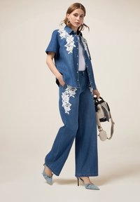 Camisa de denim y vaqueros de pierna ancha con bordado floral blanco. Color azul claro, corte relajado. La modelo lleva un bolso pequeño y zapatos azules.