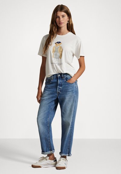 White graphic t-shirt featuring a bear design, paired with light blue loose-fit denim jeans. White sneakers complete the outfit.