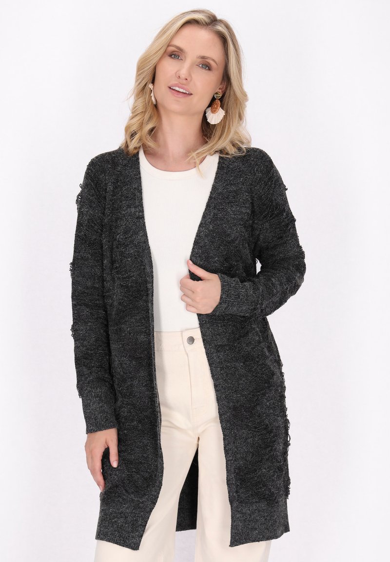 Cardigan nero a maglia con maniche lunghe, apertura frontale e dettagli testurizzati lungo i bordi. Indossato su una blusa chiara e pantaloni panna.