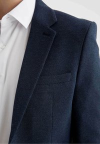 Blazer blu navy con texture e vestibilità slim, caratterizzato da un colletto a punta e una tasca sul petto discreta, indossato sopra una camicia bianca con colletto.