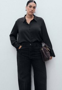 Camisa negra de satén de botones, corte relajado; combinada con jeans de mezclilla negros de cintura alta. Pequeño bolso de cuero burdeos con herrajes dorados.