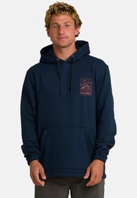 Mężczyzna z kręconymi blond włosami, ubrany w granatowy hoodie z graficznym wzorem góry i słońca na klatce piersiowej oraz ciemnymi spodniami, stojący na tle białym.