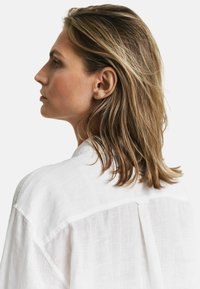 Witte linnen blouse met een losse pasvorm, voorzien van een knoopdetail op de rug. Het materiaal heeft een zachte, geweven uitstraling.
