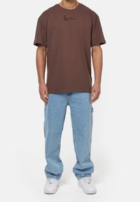 Brązowy bawełniany t-shirt z krótkim rękawem i okrągłym dekoltem. Posiada czarny logo tekstowe na piersi. Noszony z jasnoniebieskimi, luźnymi dżinsami.