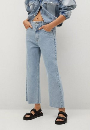 Osoba ubrana w jasnoniebieskie krótkie szerokie dżinsy, krótki dżinsowy kurtka oraz czarne pikowane sandały, stojąca na tle jednolitego tła.