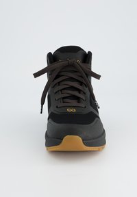 Baskets de sport noires avec une tige en maille, lacets marron foncé, semelle en caoutchouc et un accent de logo jaune. Conçues de manière structurée, avec un design à bout arrondi.