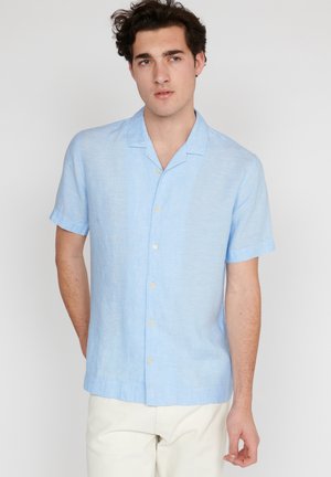 Chemise bleu clair à manches courtes et boutons, fabriquée en tissu texturé, avec un col camp et des boutons jaunes, portée avec un pantalon clair.