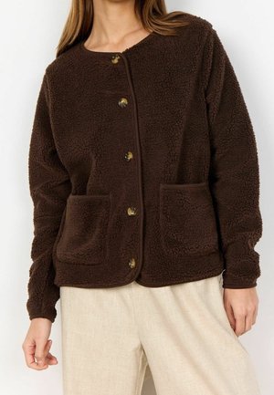 Femme portant une veste polaire marron foncé boutonnée avec deux poches avant et un pantalon beige clair, debout devant un fond uni.
