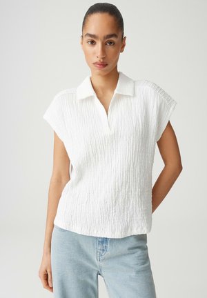 Frau mit zurückgebundenem Haar trägt ein strukturiertes weißes kurzärmeliges Hemd mit Kragen und hellblaue Jeans, stehend vor einem schlichten Hintergrund.