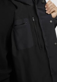 Giacca nera con tasca con zip sul lato sinistro e una texture di tessuto liscia. Ferramenta in argento con dettagli di cucitura visibili.