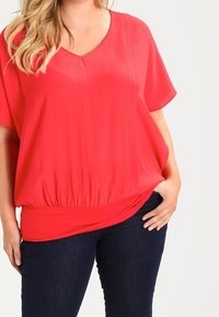 Blusa rossa, morbida e con vestibilità ampia, con scollo a V e maniche corte. La vita arricciata crea una silhouette elasticizzata. Tessuto morbido e liscio.