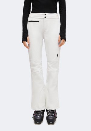 Personne portant un pantalon de ski blanc avec une poche zippée noire et des bottes de ski noires, debout devant un fond uni.