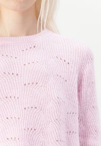 Close-up van een persoon die een lichtroze trui draagt met een tekstuurpatroon, die een gebreid ontwerp en een ronde hals laat zien.