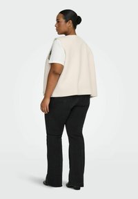 Femme avec un chignon portant une chemise blanche à manches courtes, un gilet beige sans manches, un jean noir et des chaussures noires, debout de profil sur un fond blanc.