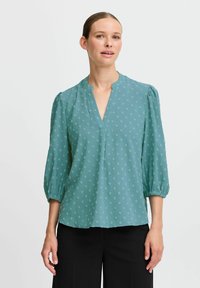 Blusa teal con scollatura a V, maniche a tre quarti a sbuffo e un motivo a pois in verde chiaro. Tessuto morbido e leggero con vestibilità rilassata.