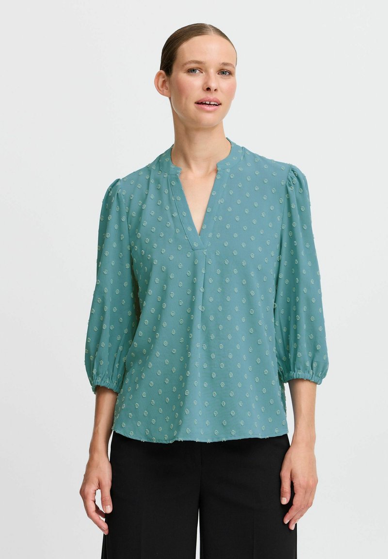 Blouse teal avec un décolleté en V, des manches bouffantes trois-quarts et un motif à pois texturé en vert clair. Tissu doux et léger avec une coupe décontractée.