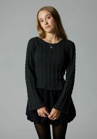 Maglione nero a maglia grossa con maniche lunghe e svasate; corpetto aderente; abbinato a una gonna nera con volant; il modello indossa anelli e una collana.