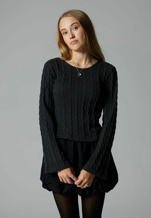 Svart kabelstickad tröja med långa, utsvängda ärmar; figurnära bodice; ihop med en volangkjol i svart; modellen bär ringar och ett halsband.