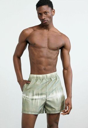 Mand uden skjorte iført grønstribede shorts med elastisk talje, poserende med den ene hånd i lommen mod en ensartet lys baggrund.