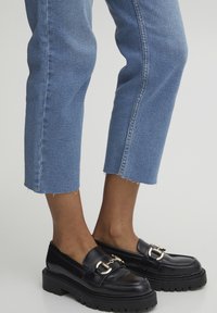 Mocasines de cuero negro con suela gruesa, con una hebilla decorativa en tono dorado. Combinados con vaqueros azules recortados con dobladillos deshilachados.