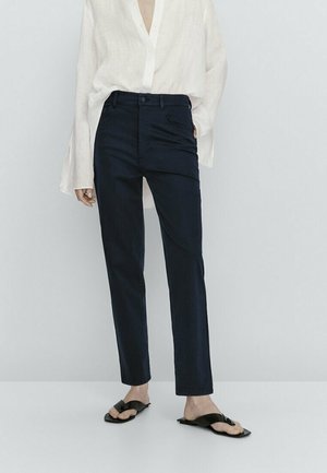 Woman wearing black straight-leg pants, white long-sleeve blouse with wide cuffs, and black flat sandals with toe loops, standing against a plain background.