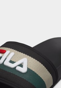 Κοντινό πλάνο μαύρης σαγιονάρας με λουράκι από μπεζ και πράσινο ριγέ ύφασμα που φέρει ανάγλυφο λευκό και κόκκινο λογότυπο "FILA".