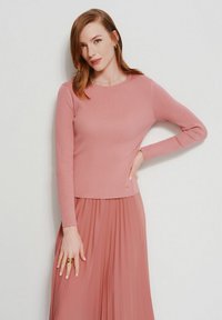 Kobieta o włosach koloru czerwonego do ramion, ubrana w dopasowany różowy sweter z prążkowanej tkaniny i plisowaną spódnicę, pozująca z ręką na biodrze na białym tle.