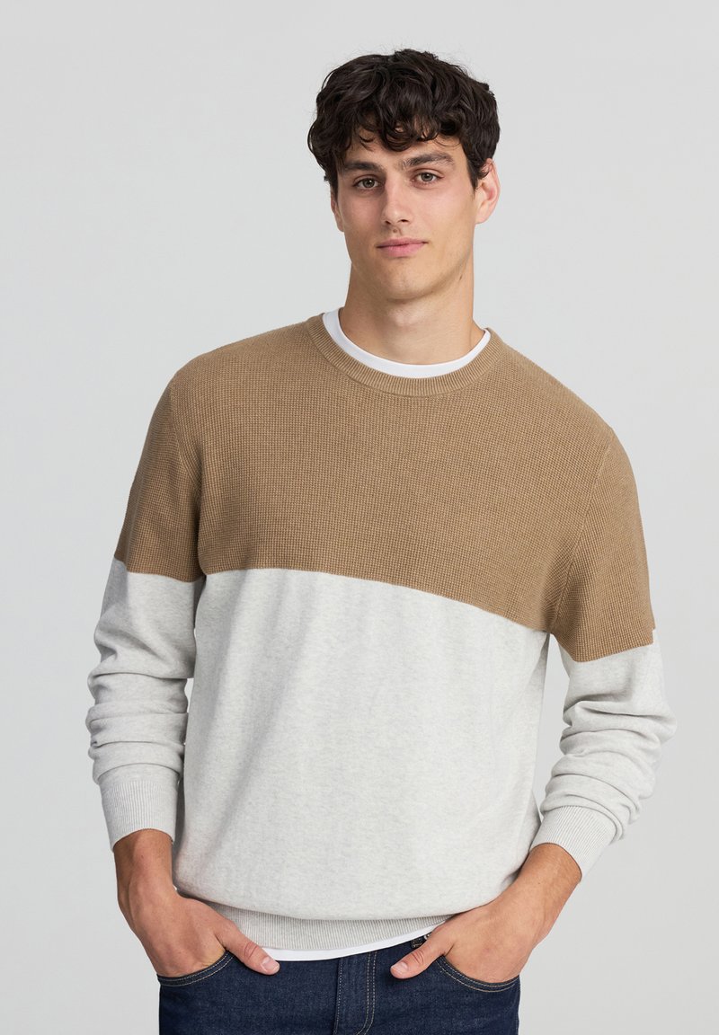 Jasnoszary sweter z teksturowaną brązową górną częścią z dzianiny. Posiada okrągły dekolt i ściągacze przy mankietach. Ręce w kieszeniach, minimalne detale.